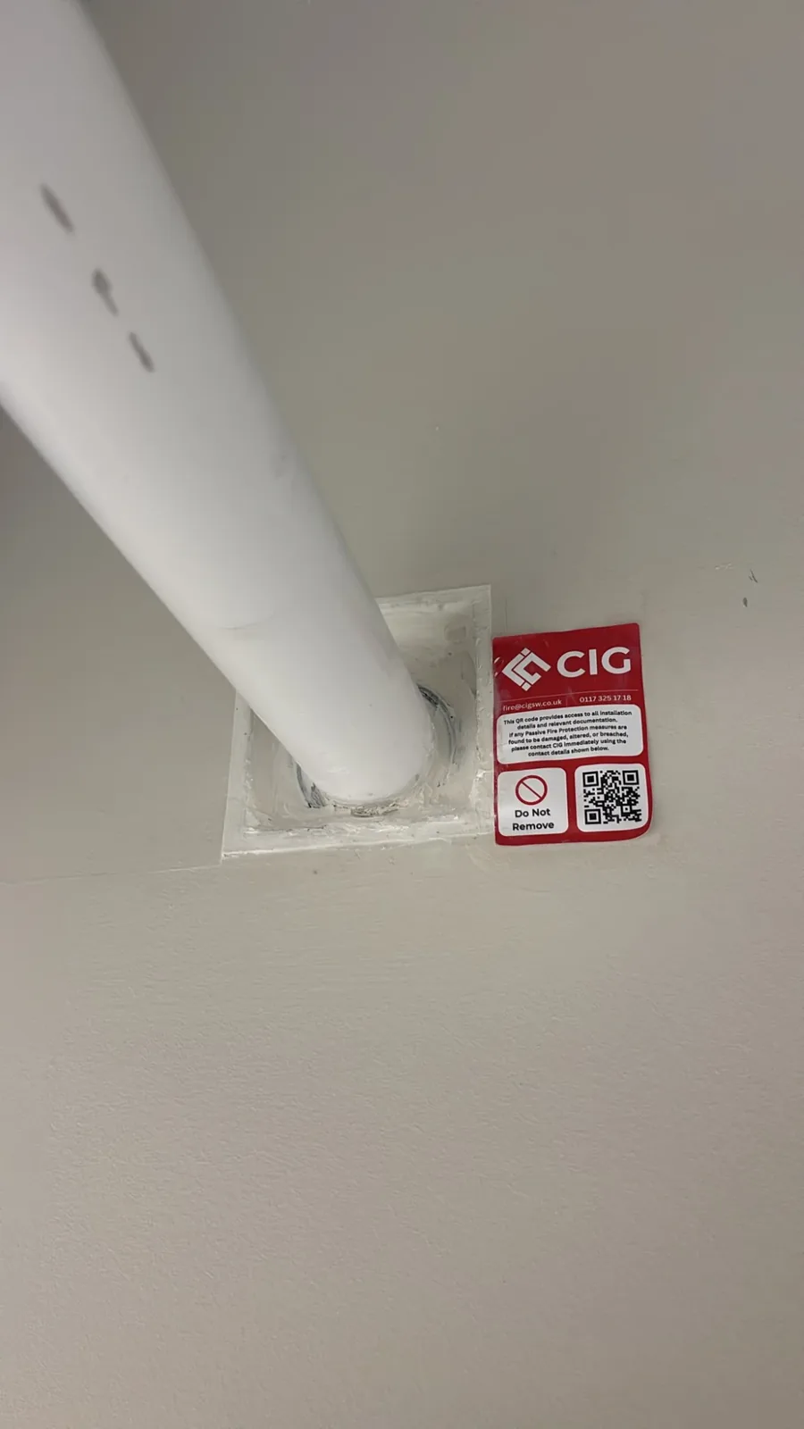 Pipe collar seal around a white plastic pipe through a wall, with a red CIG QR-coded label beside it